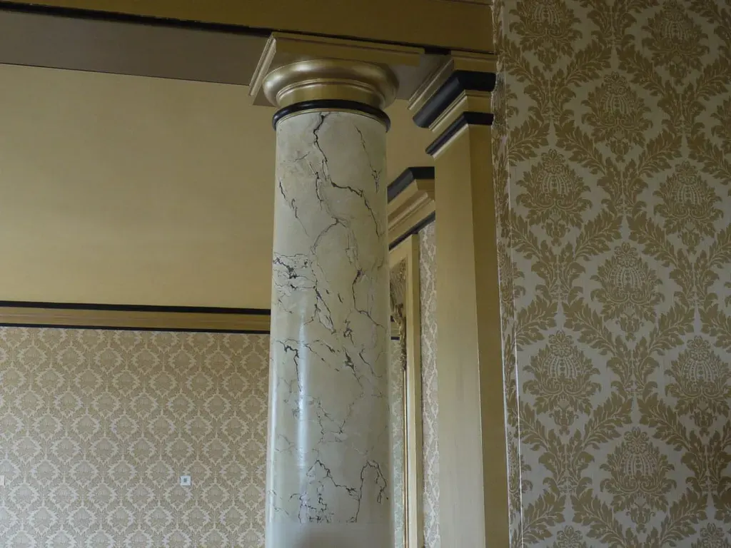 A hand-marbled column with a gilded capital, set against damask wall covering.
